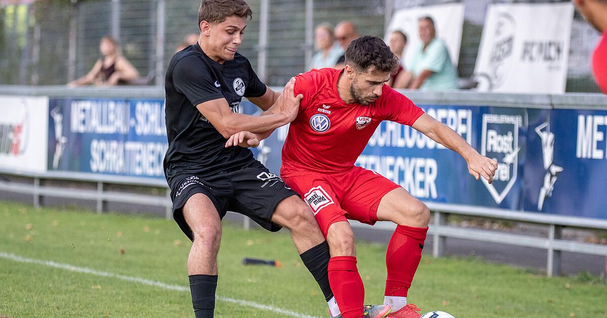 Two soccer players compete on a field. One wears a black jersey, and the other wears red. The player in red holds the ball.