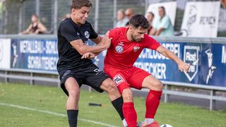 Two soccer players compete on a field. One wears a black jersey, and the other wears red. The player in red holds the ball.