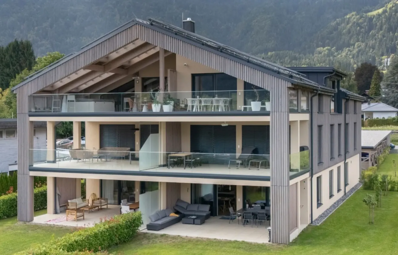 A modern, multi-story house with glass balconies and patios, surrounded by greenery. It features a kitchen, living area, and outdoor dining with a stunning mountain backdrop.
