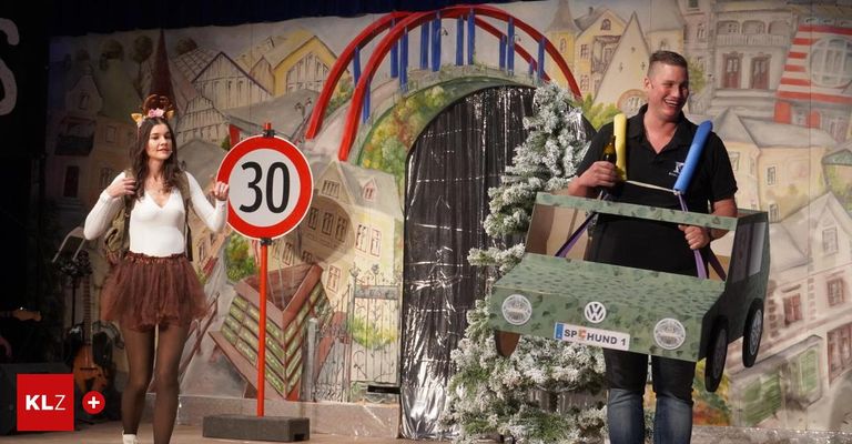 Eine Frau hält ein Schild für eine Geschwindigkeitsbegrenzung von 30 km/h, während ein Mann neben einem geschmückten Weihnachtsbaum steht. Sie befinden sich auf einer Bühne mit einer bemalten Kulisse.