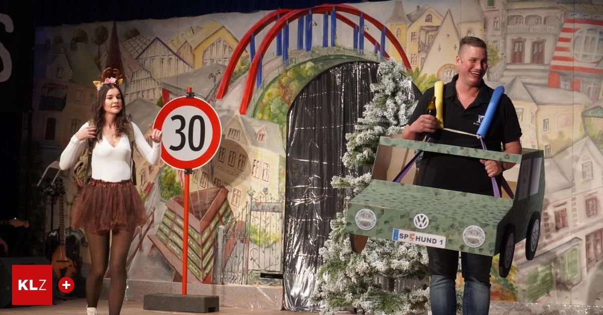 Eine Frau hält ein Schild für eine Geschwindigkeitsbegrenzung von 30 km/h, während ein Mann neben einem geschmückten Weihnachtsbaum steht. Sie befinden sich auf einer Bühne mit einer bemalten Kulisse.