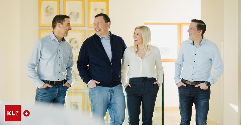 Vier Personen, zwei Männer und zwei Frauen, lächeln und stehen zusammen in einem Raum mit einer Glaswand und gerahmten Bildern an der Wand.