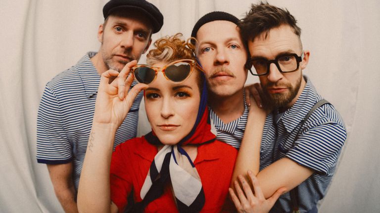 Four people pose for a photo. The woman in red wears a scarf and holds sunglasses. The man on the left wears a beanie and has a tattoo. The other two men wear glasses and striped shirts.
