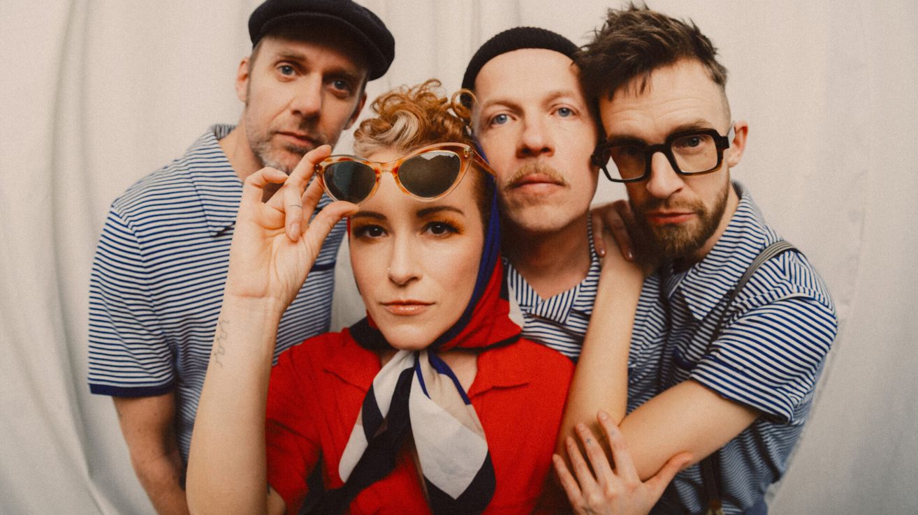 Four people pose for a photo. The woman in red wears a scarf and holds sunglasses. The man on the left wears a beanie and has a tattoo. The other two men wear glasses and striped shirts.