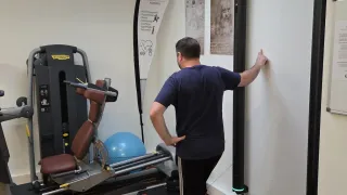 A man in a gym touching a wall with his hand while several fitness machines and equipment are behind him.