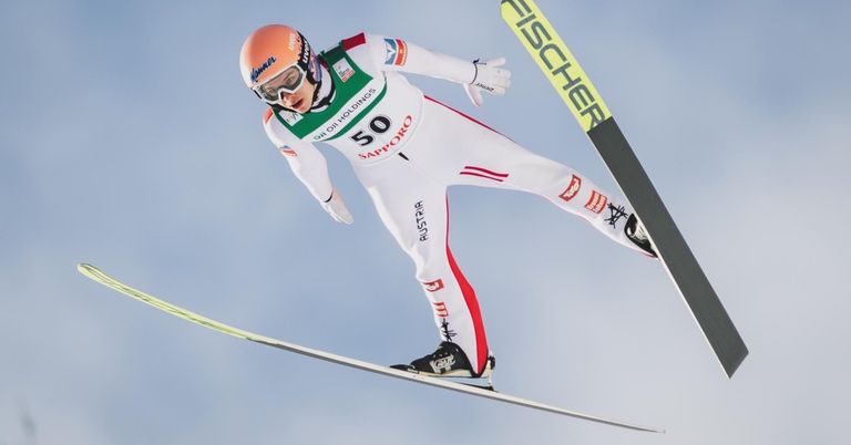 A ski jumper is mid-air, wearing a white and red ski suit with '50' on the front, an orange helmet, and goggles.