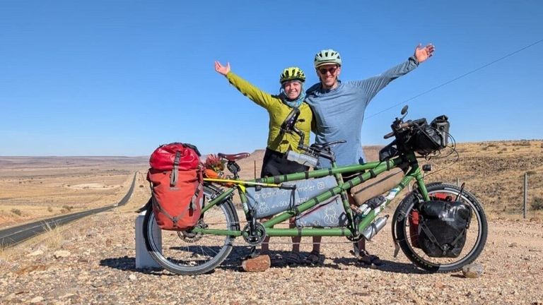 Zwei Personen stehen neben einem Tandemfahrrad, tragen Helme und lange Ärmel. Sie lächeln und heben ihre Arme. Das Fahrrad hat rote Taschen und einen grünen Rahmen.