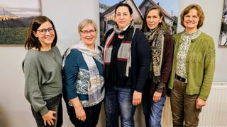 Five women stand together in front of a wall, smiling. They are all wearing scarves and some have glasses. Behind them is a photo of a house with a red roof.