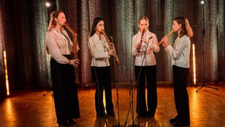 Four women in matching outfits stand on a stage, each holding a different wooden flute and playing it. They are surrounded by microphones on stands. The stage has a wooden floor and a metallic curtain backdrop.