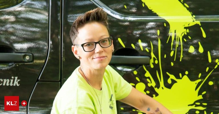 Eine Frau mit kurzen Haaren und Brille steht vor einem Fahrzeug mit schwarz-gelber Lackierung. Sie trägt ein grünes Shirt und hat Tattoos auf dem rechten Arm.