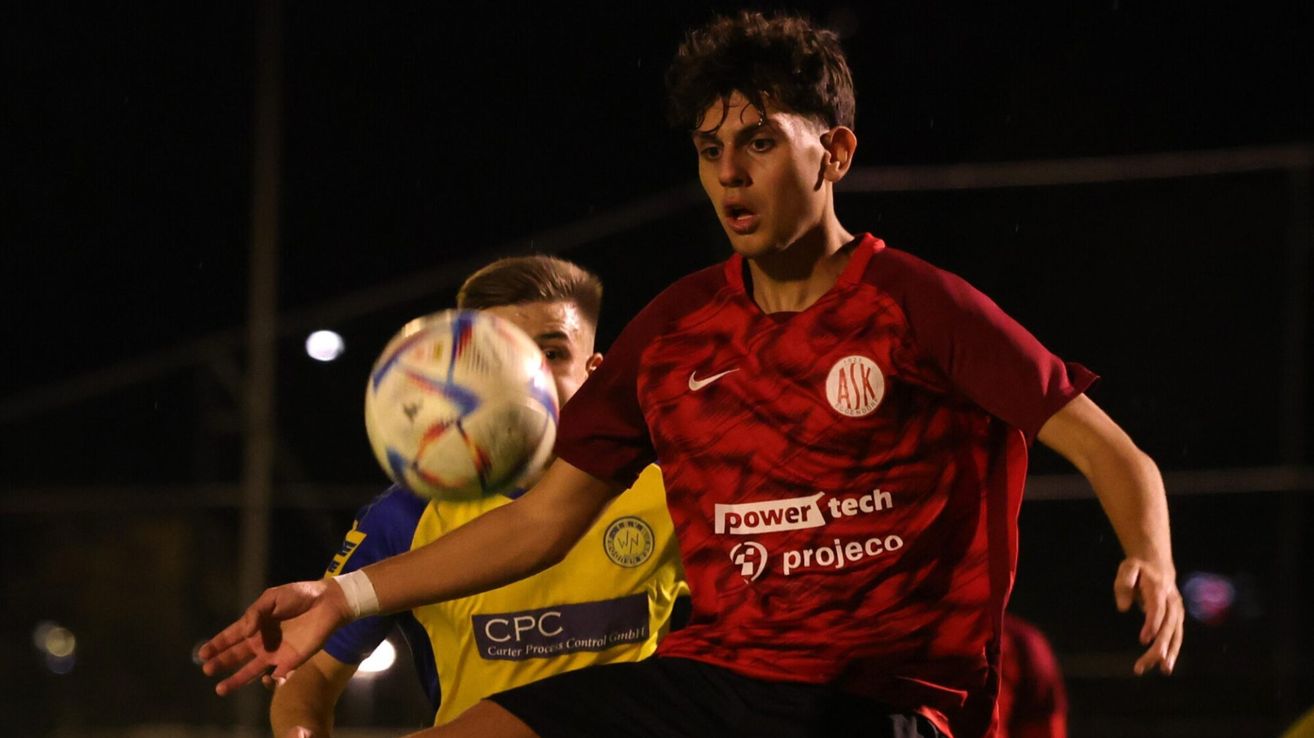 Zwei Fußballspieler spielen bei Nacht. Der Spieler in Rot streckt seinen Arm aus, um den Ball zu treffen. Der andere Spieler versucht, den Ball zu blocken.