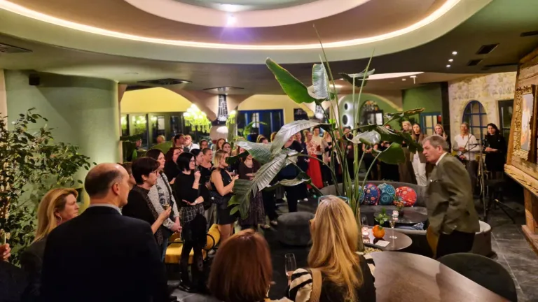 A room filled with people gathered for an event, some standing near a large plant, others sitting on chairs, with a chandelier hanging from the ceiling and various decorative items on tables.