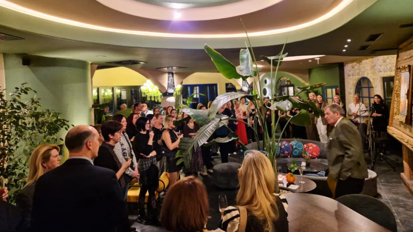 A room filled with people gathered for an event, some standing near a large plant, others sitting on chairs, with a chandelier hanging from the ceiling and various decorative items on tables.