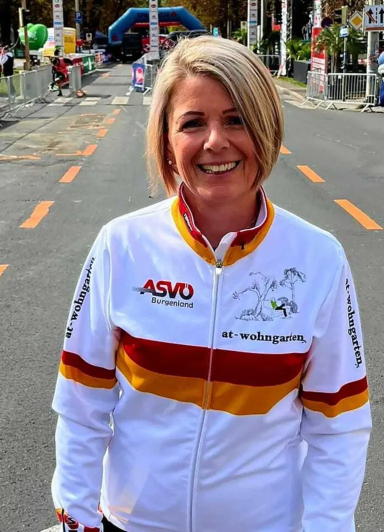 Eine Frau in einem weißen Jackett mit roten und gelben Streifen steht auf der Straße und lächelt. Das Jackett trägt die Aufschrift 'ASVO Burgenland' und 'at-wohngarten'. Hinter ihr befinden sich orange Markierungen und Metallbarrieren.