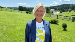 A woman with blonde hair, wearing a blue blazer and a lemon-printed t-shirt, is standing in a grassy field. Behind her, there are houses, trees, and mountains.