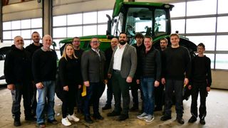 A group of individuals, both men and women, are posing for a photograph in a garage, with a John Deere tractor in the background.
