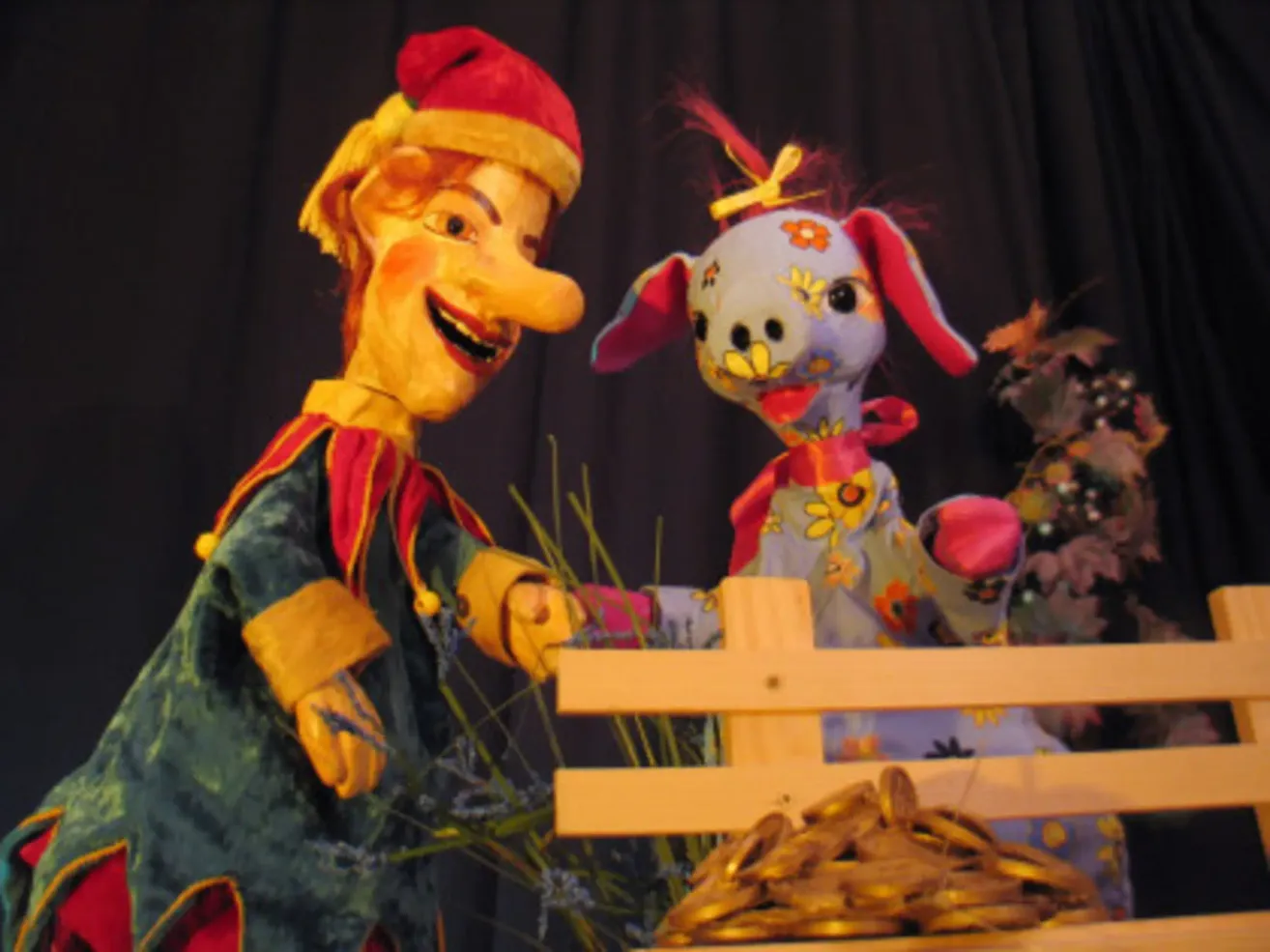 Two puppets, one dressed as a jester, are standing by a wooden fence with a pile of gold coins in front of them.