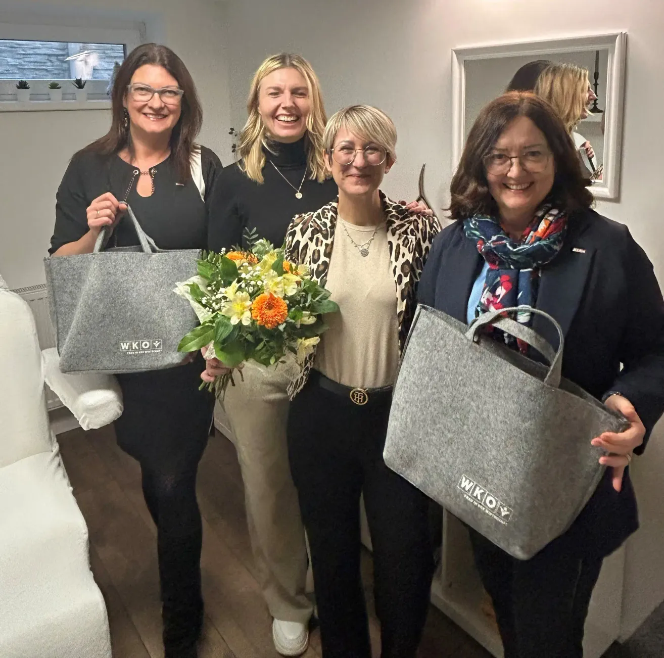 Fünf Frauen stehen zusammen und halten Taschen, eine hält einen Blumenstrauß. Alle lächeln, tragen eine Brille und sind lässig gekleidet. Hinter ihnen befindet sich ein Spiegel mit der Reflexion eines Raumes.