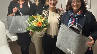 Five women stand together holding bags, one holding a bouquet of flowers. All are smiling, wearing glasses, and dressed casually. Behind them is a mirror with a reflection of a room.