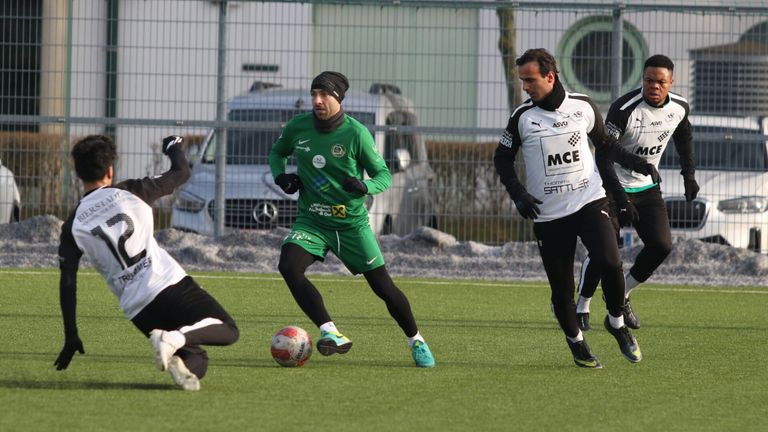 Zwei Fußballspieler sind auf einem Feld, einer in Grün und der andere in Weiß. Sie rennen und jagen einem Ball hinterher. Dahinter ist ein Zaun und geparkte Autos.