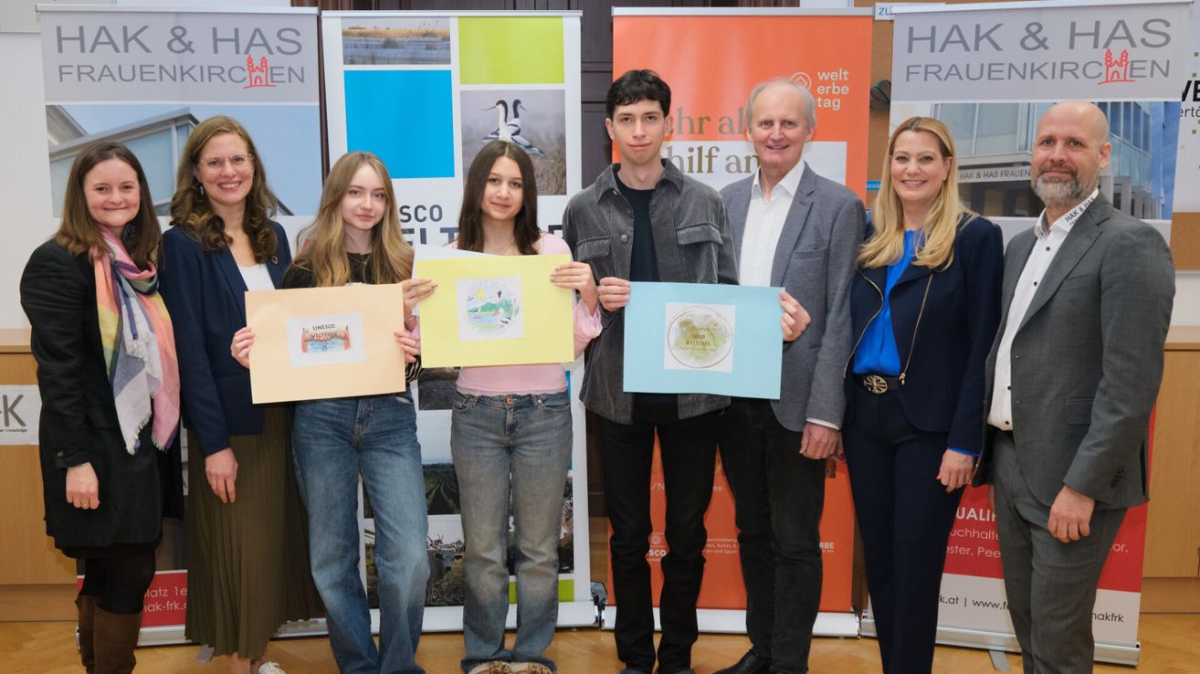 Fünf Personen stehen zusammen und halten gerahmte Zeichnungen. Zwei Frauen halten gelbe und blaue Zeichnungen, und drei Männer halten blaue Zeichnungen. Sie stehen vor Bannern.