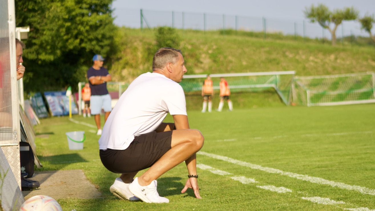 An adult male football coach is crouching on the grass, possibly giving instructions to his team. Behind him, players are standing and watching. In the background, there is a fence and a blurry view of a grassy hill.