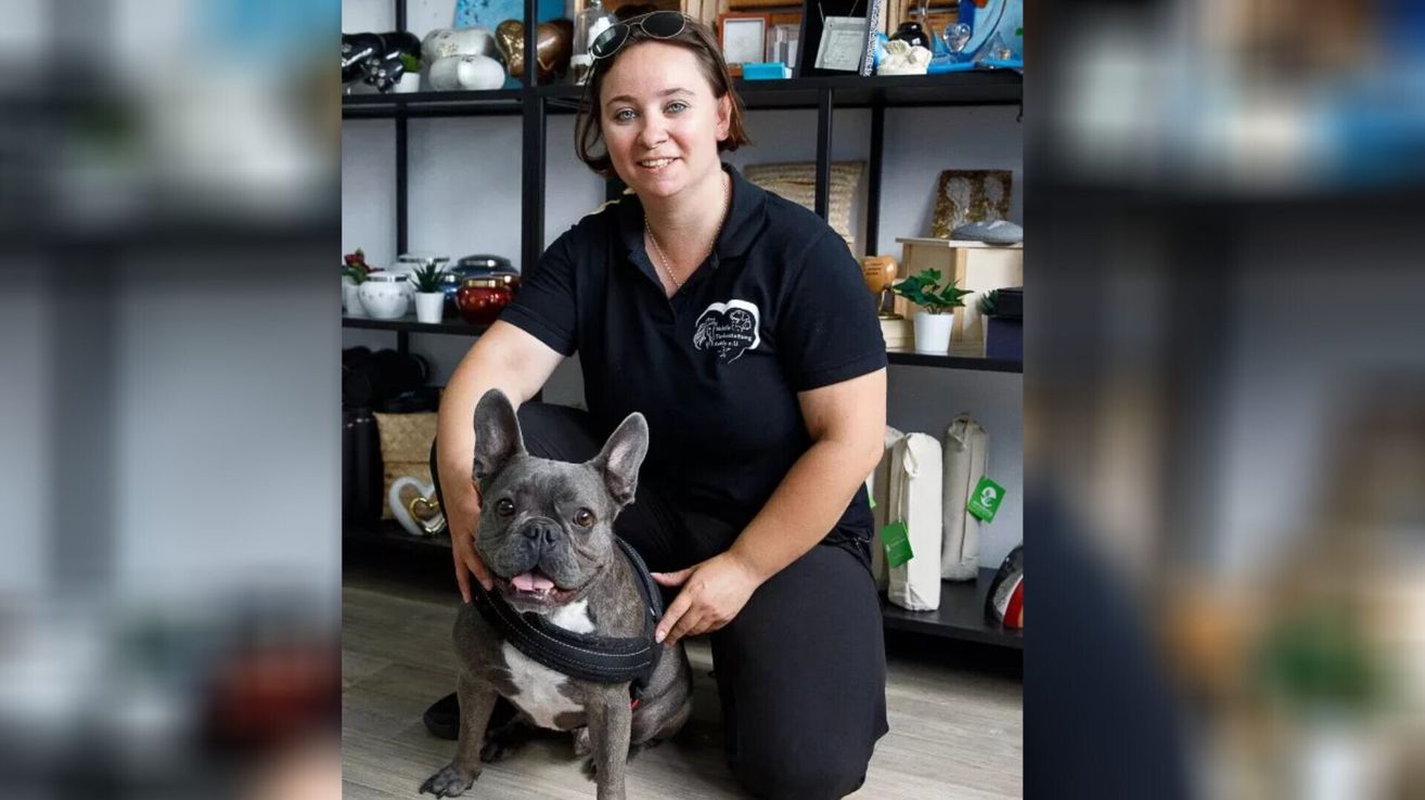 Eine Frau in schwarzer Uniform kniet lächelnd neben einem französischen Bulldoggen-Welpen mit Geschirr. Hinter ihr steht ein Regal mit verschiedenen Gegenständen.