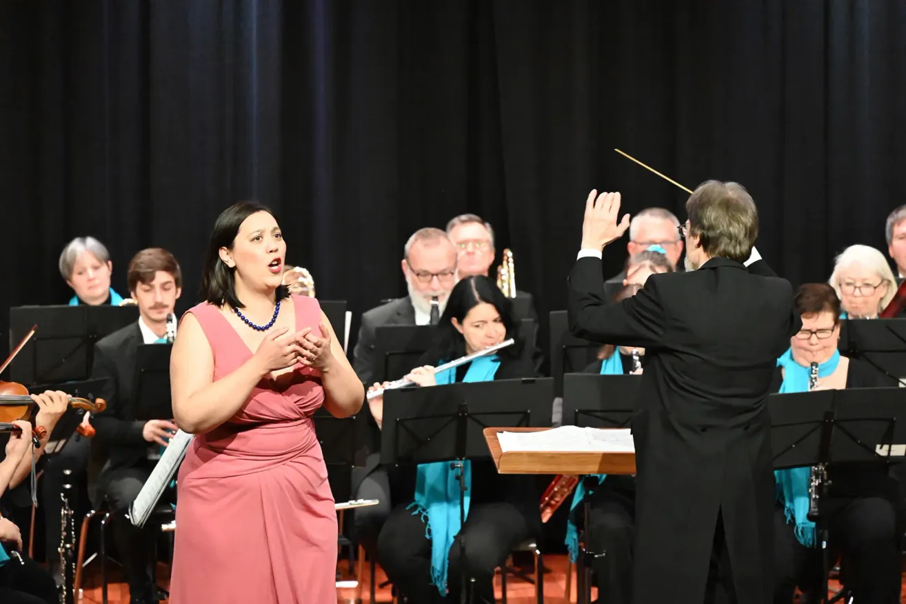 Eine Sängerin in einem rosa Kleid singt, während das Orchester hinter ihr spielt. Der Dirigent dirigiert die Aufführung. Die Orchestermitglieder sitzen vor Notenständern.