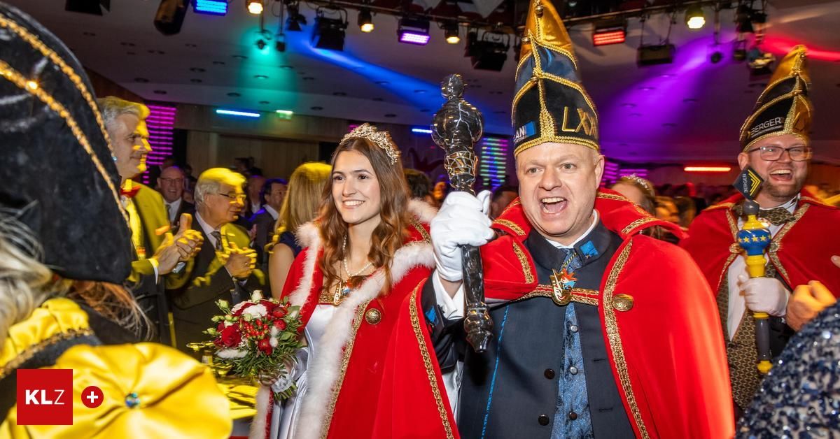Ein Mann und eine Frau in roten und goldenen Kostümen stehen in einem festlichen Saal mit vielen Gästen, der Mann hält eine Trophäe und einen Blumenstrauß.