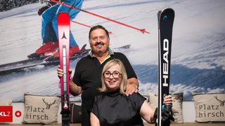 Ein Mann und eine Frau stehen nebeneinander und halten Skier. Der Mann trägt ein schwarzes Poloshirt, und die Frau trägt ein schwarzes Kleid. Sie lächeln und scheinen für ein Foto zu posieren.