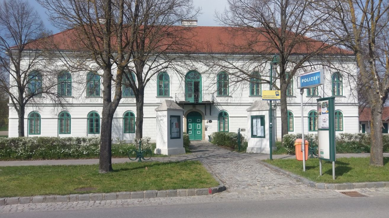 Ein weißes Gebäude mit rotem Dach, grünen Fenstern und einer grünen Tür befindet sich in einem Park. Es gibt zwei weiße Säulen auf jeder Seite des Eingangs.