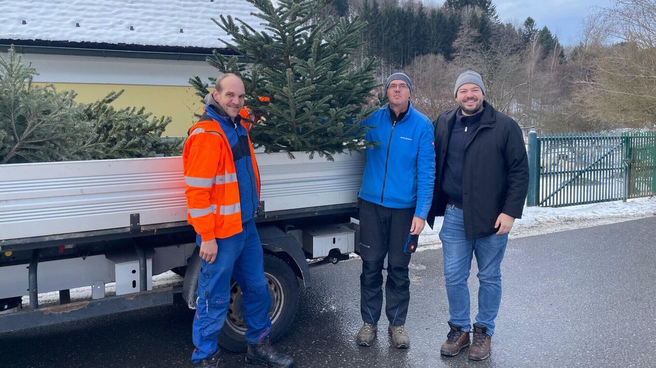 Drei Männer stehen neben einem mit Weihnachtsbäumen beladenen Lastwagen und lächeln. Der Mann links trägt einen orangefarbenen Sicherheitsoverall, der Mann in der Mitte eine blaue Jacke und der Mann rechts eine schwarze Jacke. Hinter ihnen befindet sich eine verschneite Landschaft.