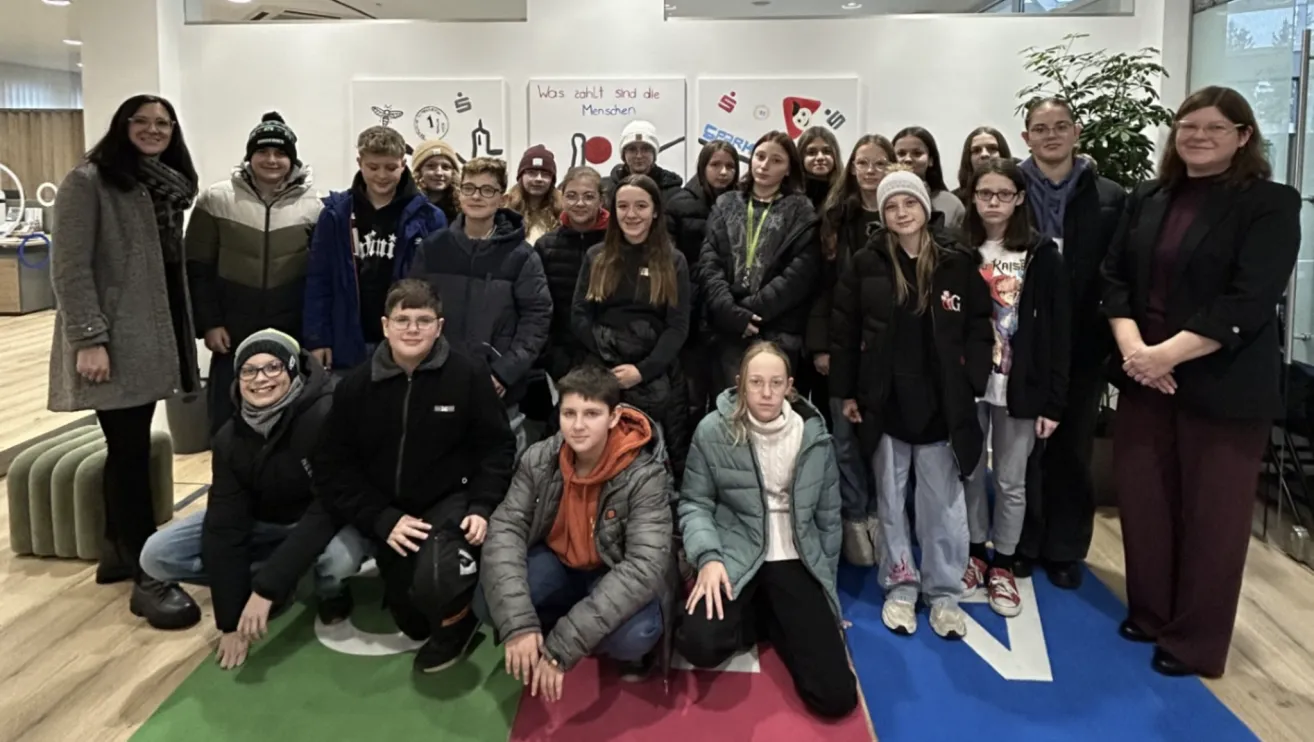 Eine Gruppe von Schülern posiert für ein Foto vor einer Wand mit verschiedenen Plakaten, darunter eines mit der Aufschrift 'Was zählt sind die Menschen'.