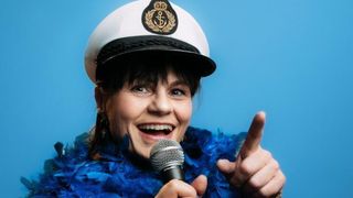 Eine Frau in einer Kapitänsmütze lächelt und hält ein Mikrofon. Sie zeigt den Daumen nach oben und trägt eine blaue Federboa.