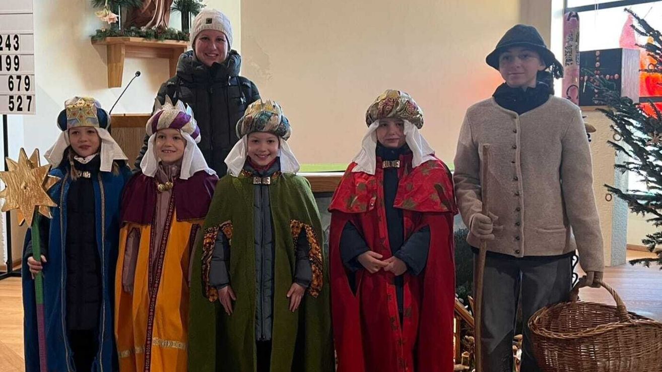 Fünf Kinder und eine erwachsene Frau in mittelalterlichen Kostümen posieren für ein Foto. Die Kinder tragen farbenfrohe Roben und Hüte, während die erwachsene Frau eine schwarze Jacke trägt. Hinter ihnen ist eine schlichte weiße Wand.