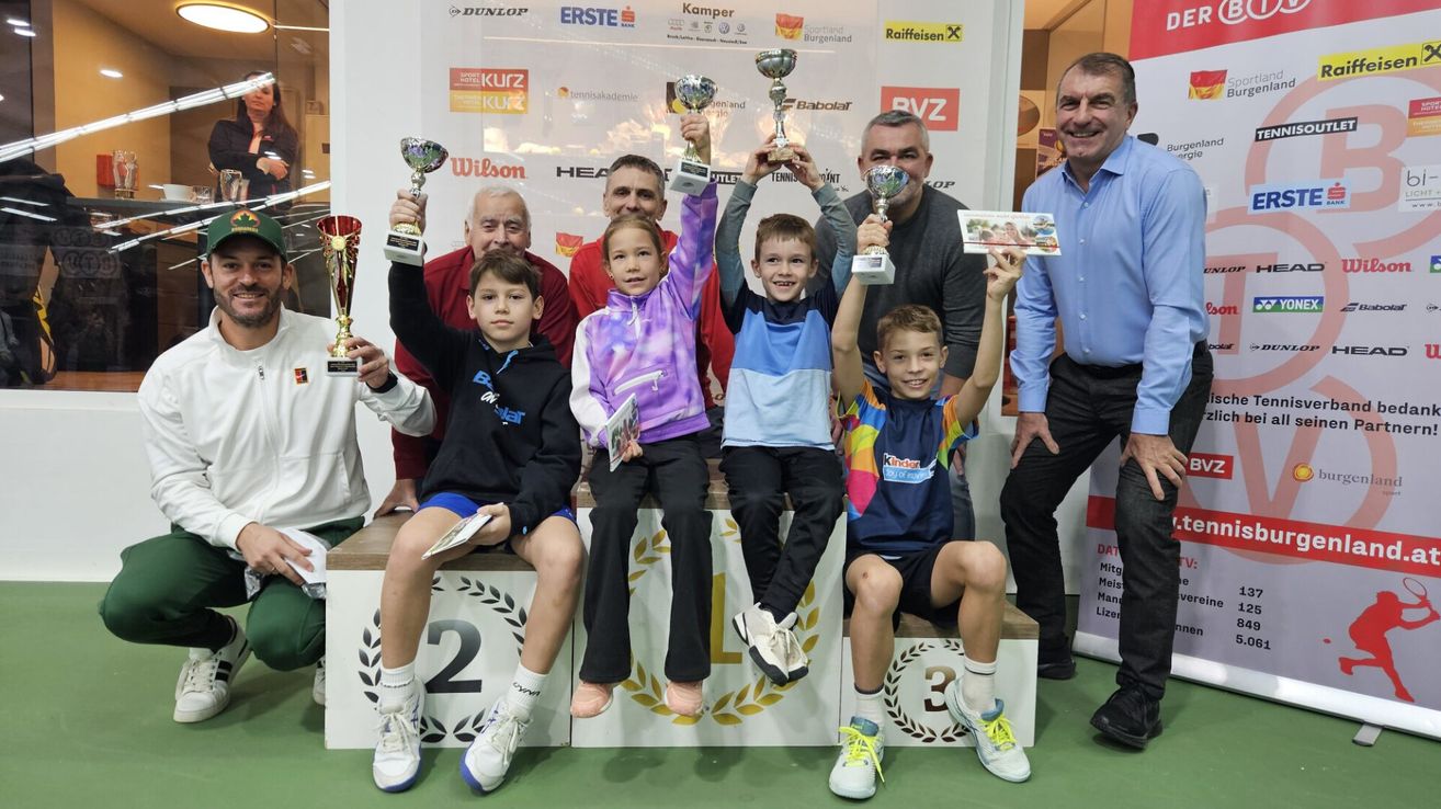 Fünf Kinder und zwei Erwachsene stehen mit Trophäen in den Händen auf einem Podium. Sie lächeln und scheinen sich bei einer Sportveranstaltung zu befinden. Hinter ihnen hat eine Wand verschiedene Werbungen.