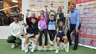 Fünf Kinder und zwei Erwachsene stehen mit Trophäen in den Händen auf einem Podium. Sie lächeln und scheinen sich bei einer Sportveranstaltung zu befinden. Hinter ihnen hat eine Wand verschiedene Werbungen.