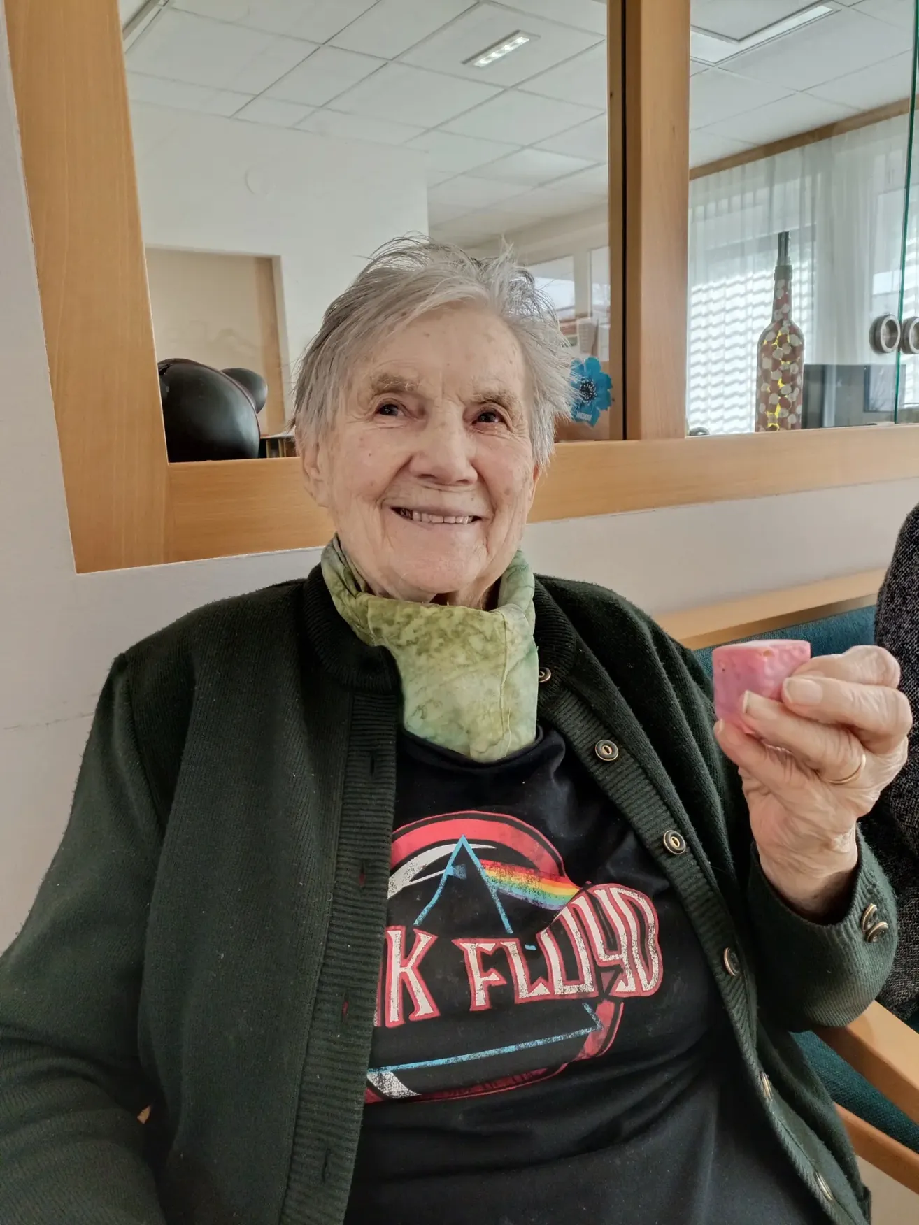 Eine ältere Frau mit grünem Schal lächelt und hält ein rosa Objekt in der Hand, sitzt in einem Raum mit Spiegel und Fenster dahinter.