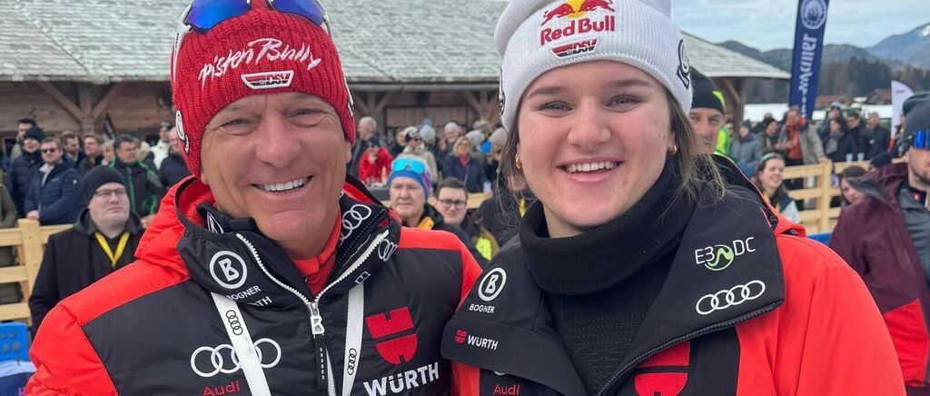 Ein Mann und eine Frau in roten und schwarzen Skijacken lächeln in die Kamera und tragen Wintermützen. Der Mann hat einen roten Hut mit Logo und Sonnenbrille obenauf. Sie stehen vor einem verschwommenen Publikum.