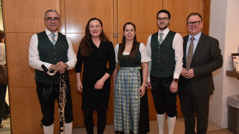 Fünf Personen, drei Männer und zwei Frauen, in traditioneller Kleidung, posieren für ein Foto. Ein Mann auf der linken Seite hält ein Musikinstrument. Sie stehen vor einer Holzwand.