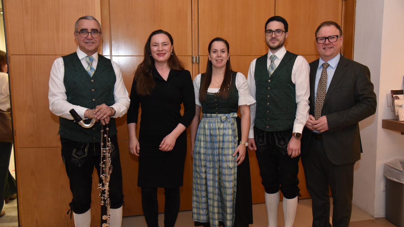 Fünf Personen, drei Männer und zwei Frauen, in traditioneller Kleidung, posieren für ein Foto. Ein Mann auf der linken Seite hält ein Musikinstrument. Sie stehen vor einer Holzwand.
