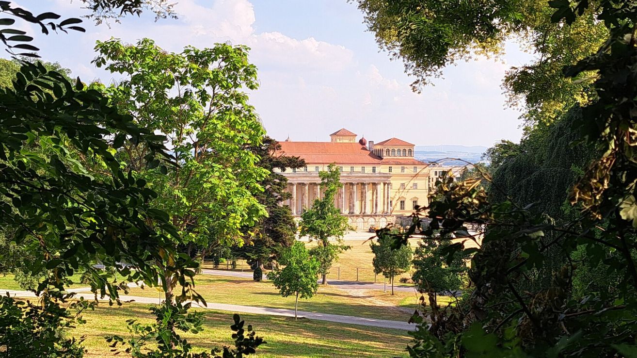 Ein klassisches Gebäude mit rotem Dach ist von üppigen Bäumen in einem Park umgeben. Das Gebäude hat viele Fenster und Säulen. Der Himmel ist teilweise bewölkt.