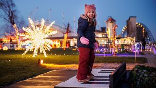 Ein junges Mädchen steht lächelnd auf einer klavierartigen Plattform und trägt eine Winterkleidung vor einer Weihnachtsbeleuchtung.