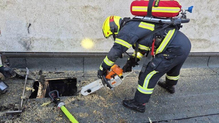 Ein Feuerwehrmann in gelbem Helm und Schutzausrüstung benutzt eine Kettensäge auf einem Dach. Die Kettensäge schneidet durch Schutt. Eine Schläuche ist in der Nähe.