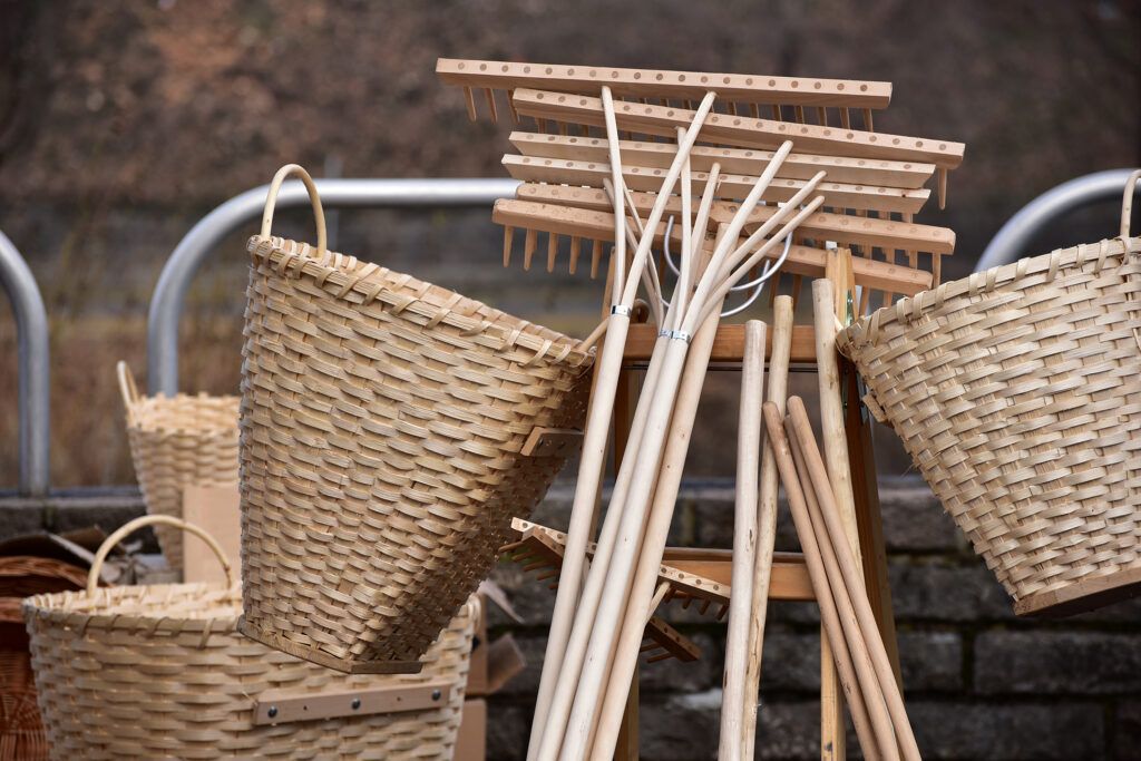Eine Sammlung von Weidenkörben und Holzwerkzeugen, einschließlich Rechen und Kämme, wird auf einem Holzständer vor einer Steinmauer ausgestellt.