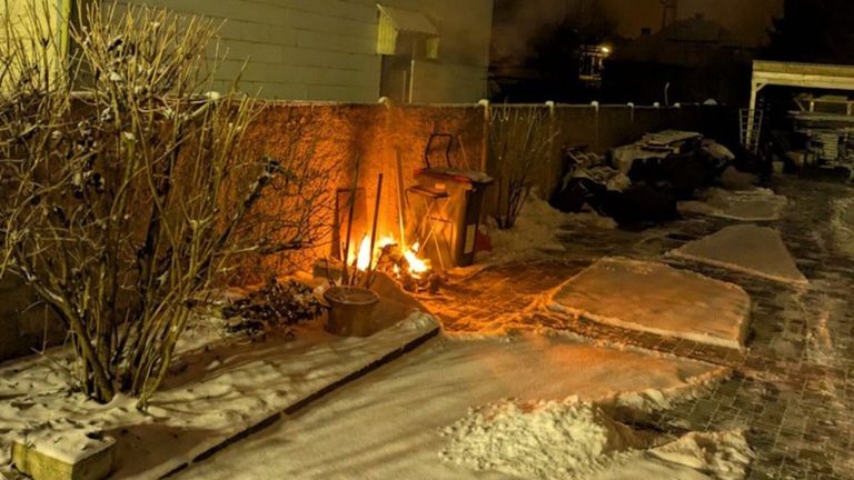 Eine Szene eines Hinterhofs bei Nacht zeigt ein Feuer mit Flammen, umgeben von einer Ziegelmauer. Ein Mülleimer ist in der Nähe. Schnee bedeckt den Boden und die Pflanzen.