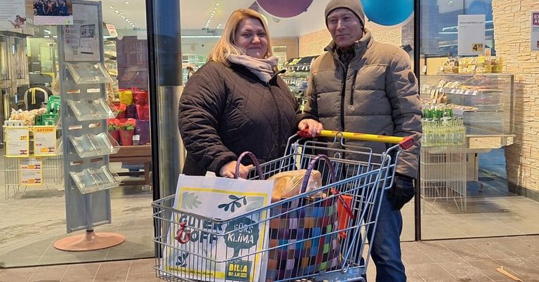 Ein Mann und eine Frau stehen in einem Geschäft mit einem vollen Einkaufswagen und lächeln in die Kamera. Hinter ihnen sind Regale mit Waren und Luftballons zu sehen.