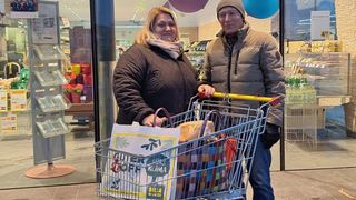 Ein Mann und eine Frau stehen in einem Geschäft mit einem vollen Einkaufswagen und lächeln in die Kamera. Hinter ihnen sind Regale mit Waren und Luftballons zu sehen.