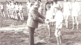 Ein Schwarz-Weiß-Foto zeigt eine Gruppe junger Athleten auf einem Rasenfeld. Ein älterer Mann in einem Anzug überreicht einer jungen Mädchen eine Auszeichnung, die lächelt. Dahinter stehen weitere Athleten in einer Reihe.