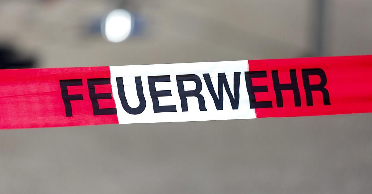 Ein rot-weißes Polizeiband mit schwarzem Text 'FEUERWEH' vor grauem Hintergrund mit schwacher weißer Lichtquelle.